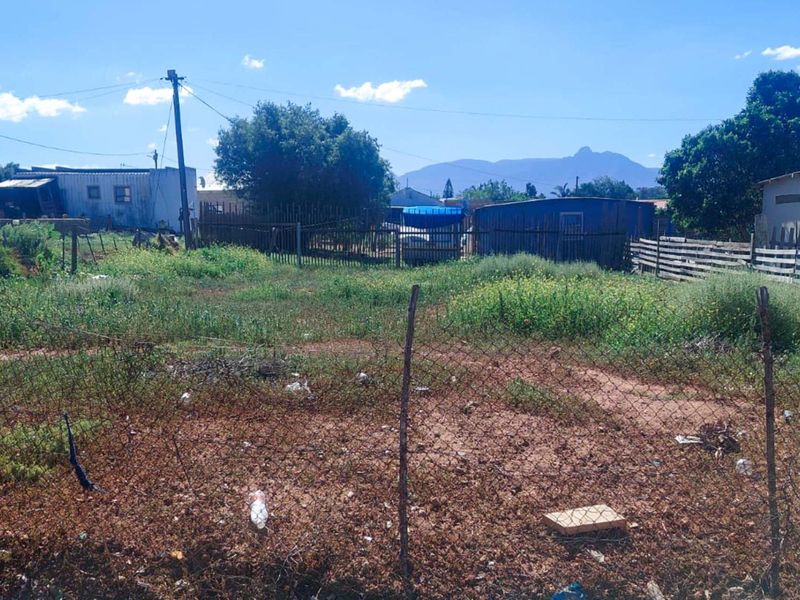 A vacant residential plot featuring wire mesh fencing and a utility pole, with surrounding neighborhood structures and mountain views in the distance.
