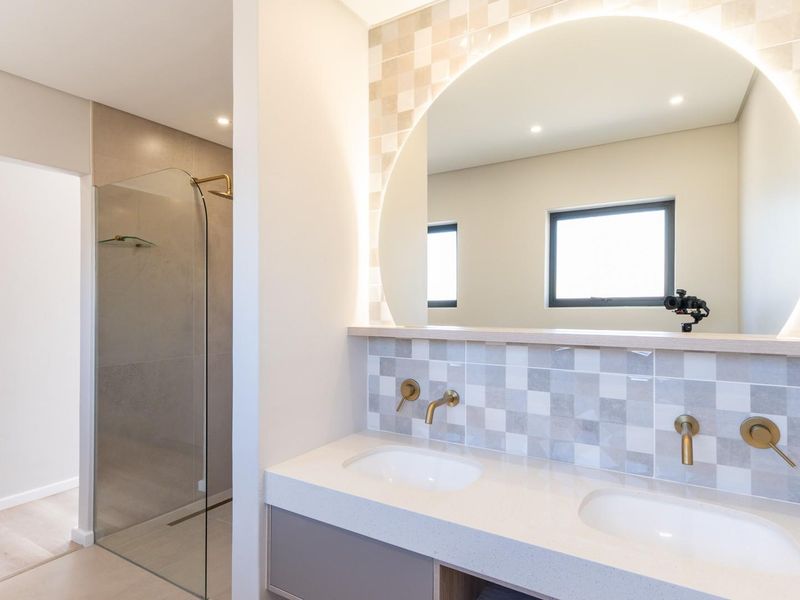Modern bathroom featuring a double vanity with stone countertop, wall-mounted brass faucets, a large backlit circular mirror, and a glass-enclosed walk-in shower.