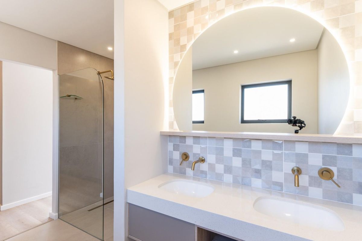 Modern bathroom featuring a double vanity with stone countertop, wall-mounted brass faucets, a large backlit circular mirror, and a glass-enclosed walk-in shower.