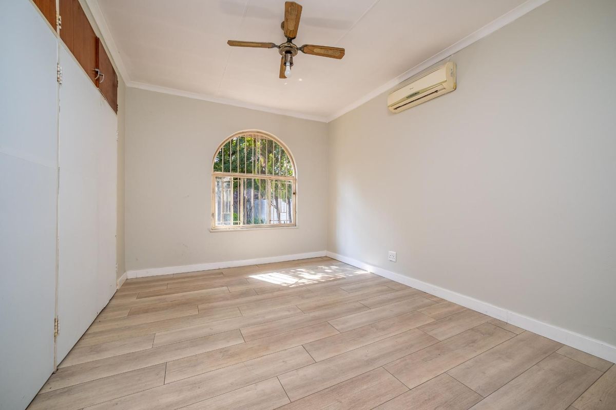 A bright bedroom featuring light wood-look flooring, a ceiling fan, a wall-mounted air conditioning unit, and an arched window with security bars.
