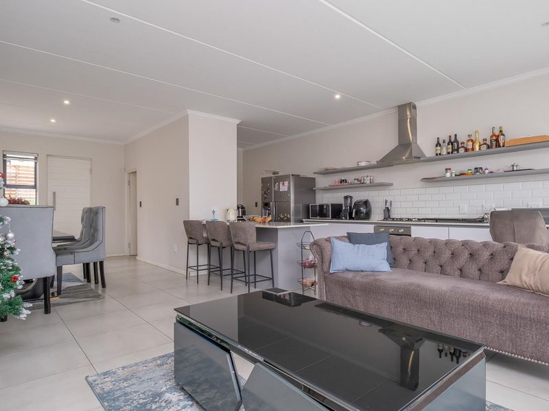 A modern open-plan living and kitchen area featuring a central island, subway tile backsplash, floating shelving, and neutral tiled flooring throughout.