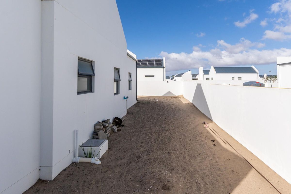 A side exterior view of the property featuring a white rendered wall, window openings, and an unlandscaped sandy yard area.