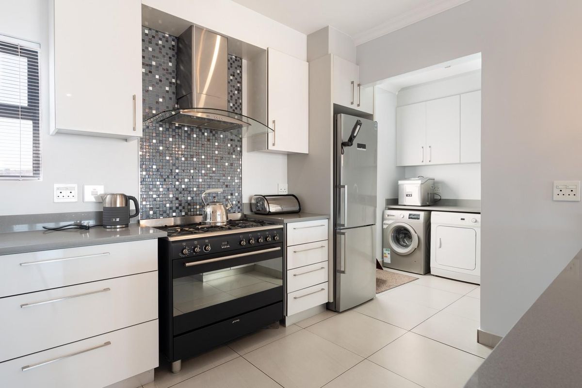 Modern kitchen featuring white cabinetry, stone countertops, a mosaic tile backsplash, and a stainless steel extractor fan, with an adjoining scullery area.