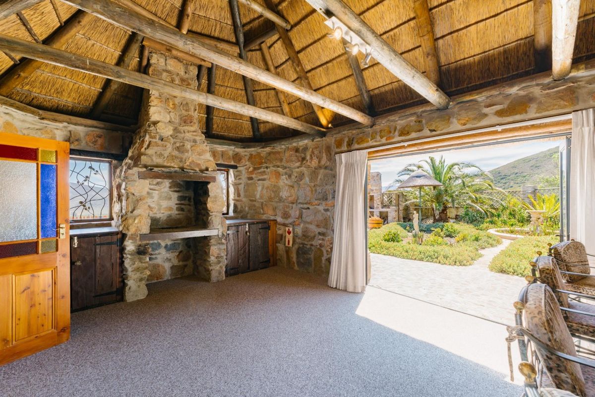A rustic entertainment area featuring a stone fireplace, exposed timber beams, and a thatched roof, opening directly onto a landscaped garden patio.
