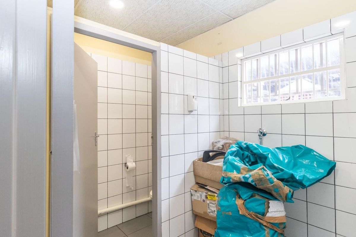 A commercial restroom facility featuring white wall tiling, a drop ceiling, and a separate toilet cubicle with a security-barred window.