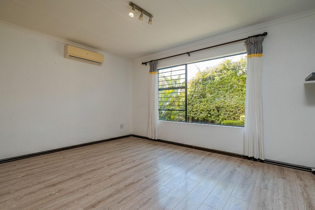 A bright, empty bedroom featuring wood-look laminate flooring, a wall-mounted air conditioning unit, and a large window overlooking the garden.
