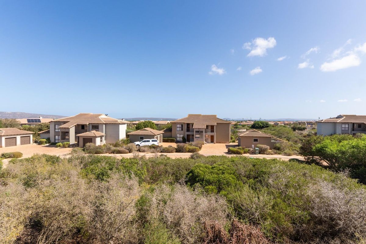 An elevated aerial view of a residential apartment complex featuring multiple buildings with tiled roofs, surrounded by natural coastal vegetation and gravel access paths.