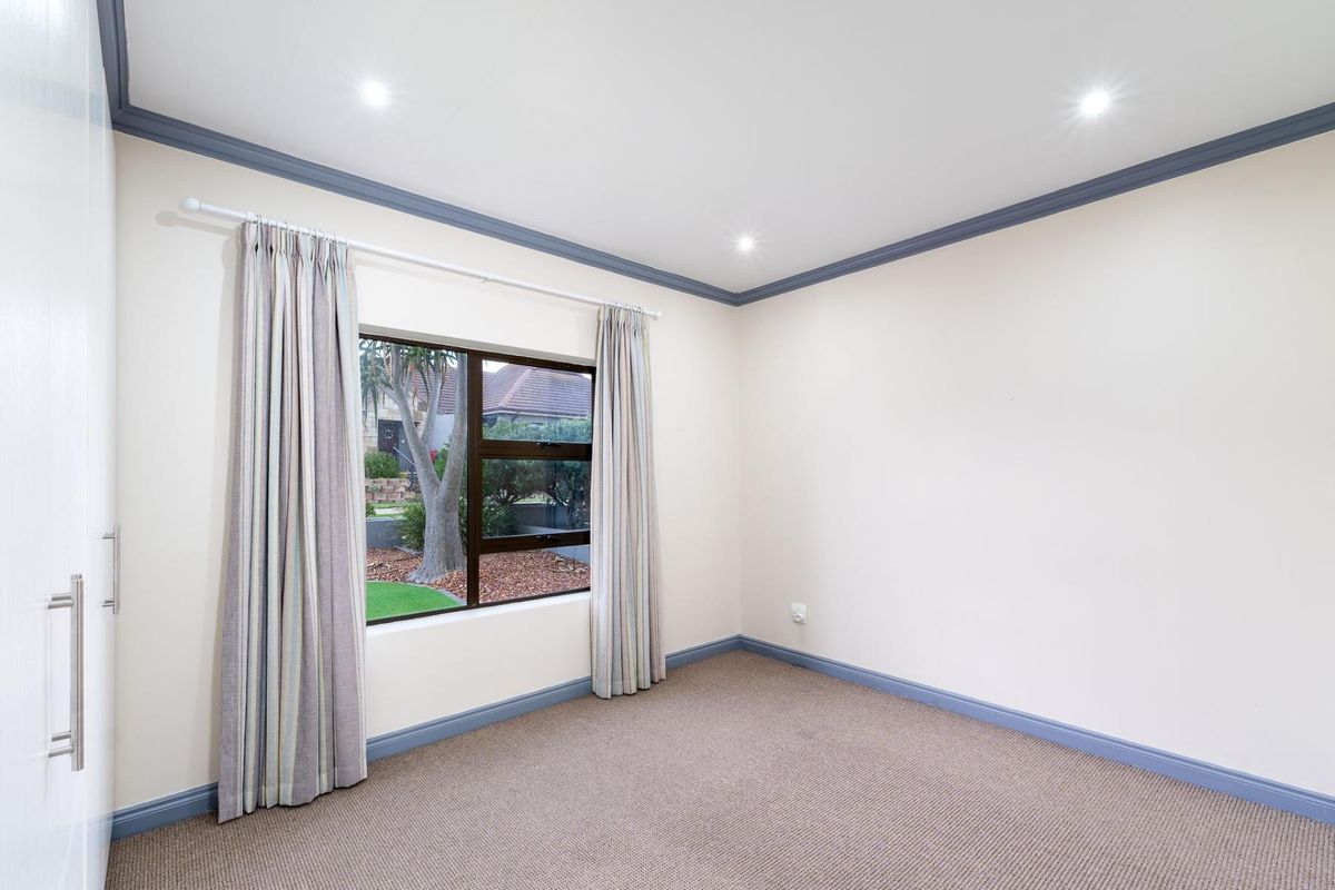 A bright, neutral-toned bedroom featuring built-in cupboards, carpeted flooring, and a window overlooking the garden.
