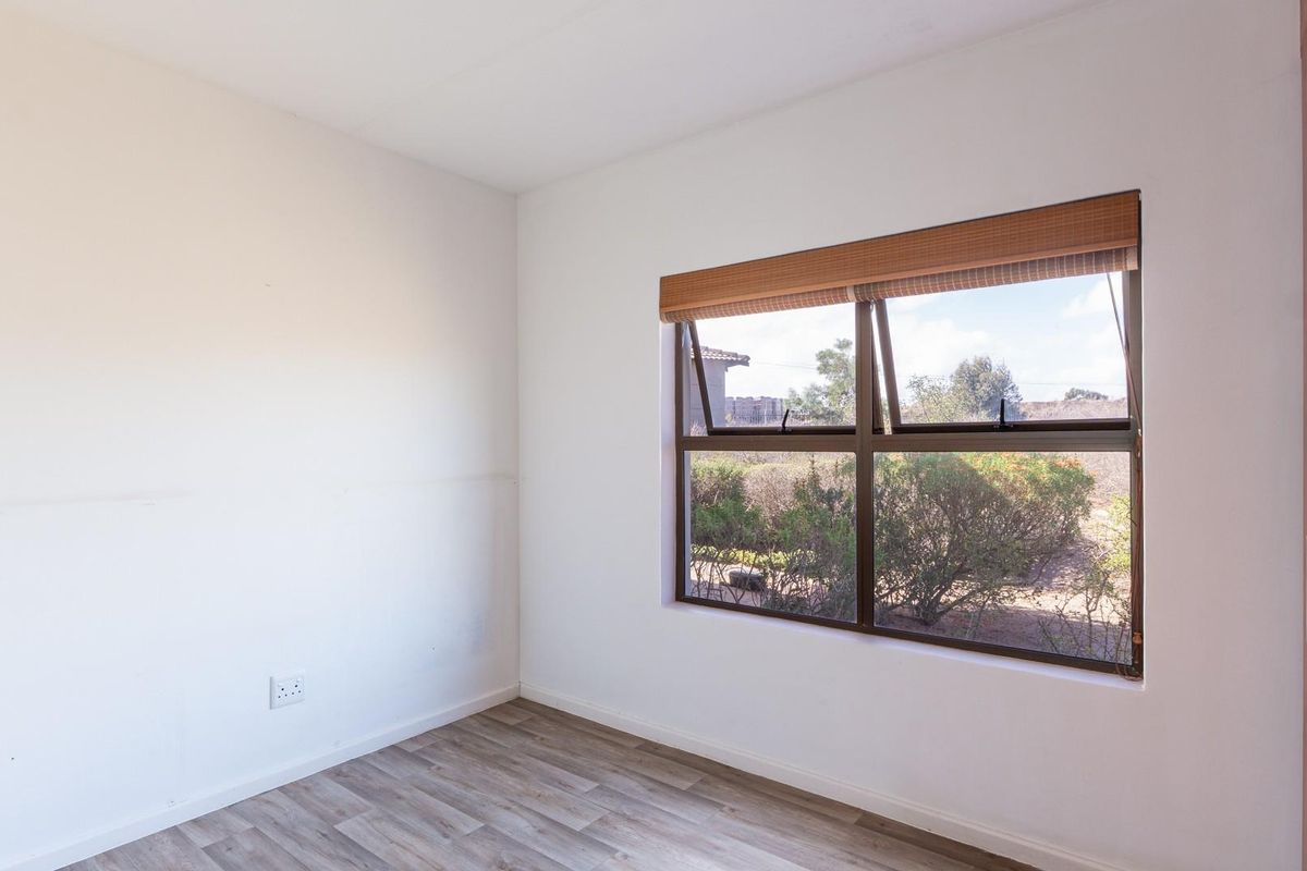 A bright, empty bedroom featuring wood-look laminate flooring, a built-in wardrobe, and a window with a roller blind.