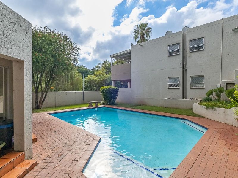 Communal swimming pool area featuring brick paving, landscaped garden borders, and views of the surrounding townhouse complex architecture.