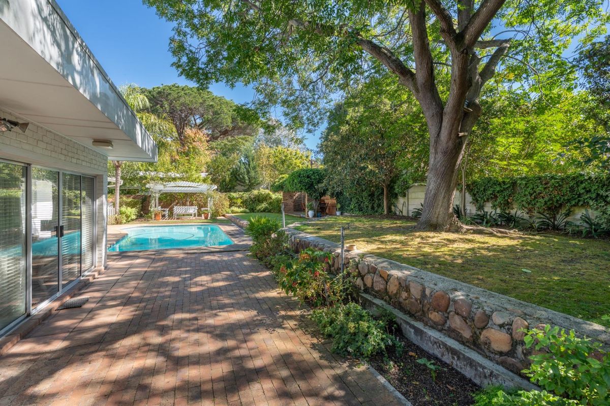 A spacious rear garden featuring a swimming pool, brick-paved patio area, mature trees, and a stone retaining wall.
