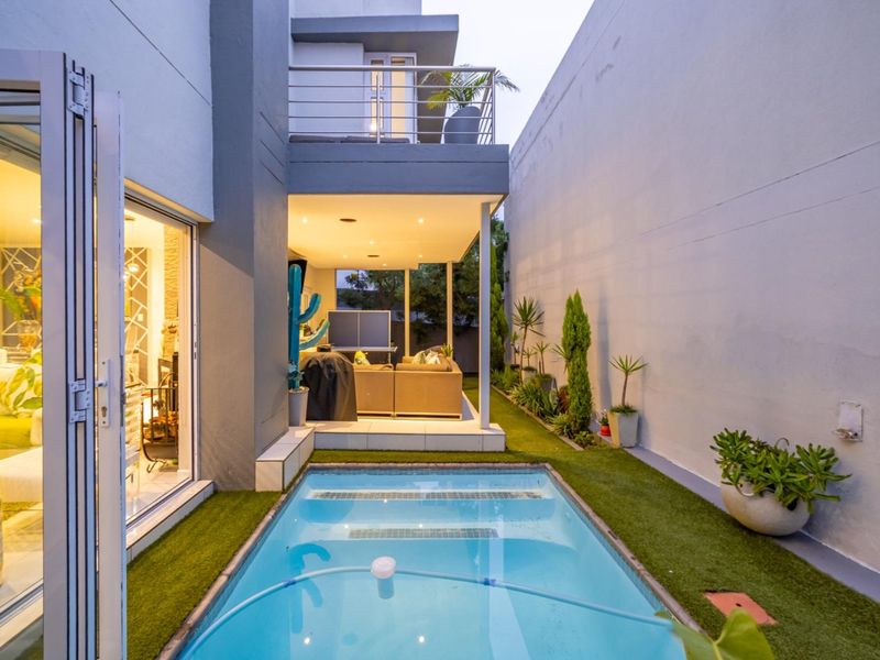 A modern townhouse rear exterior featuring a compact swimming pool, artificial lawn, and a covered patio area with recessed lighting.