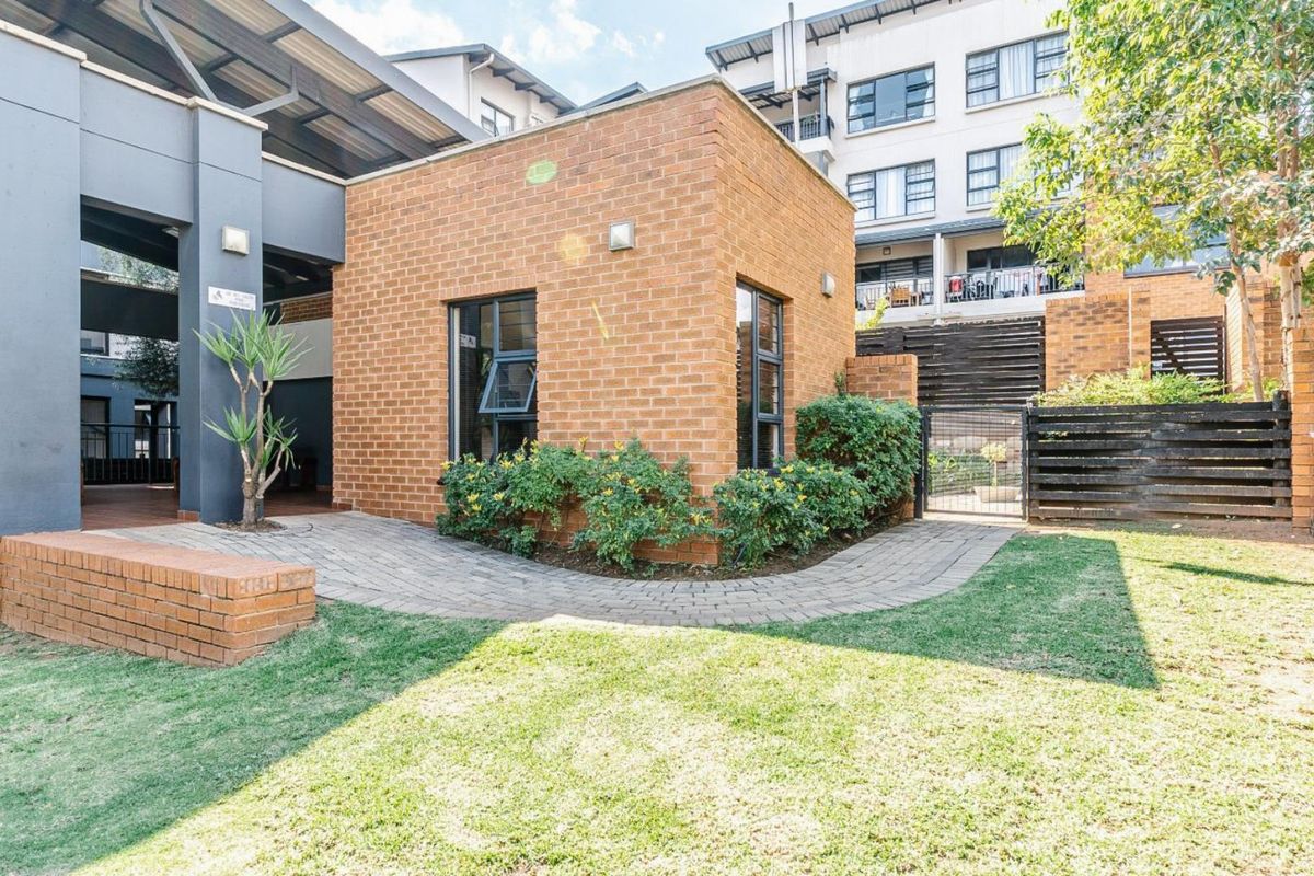 A view of the apartment complex exterior featuring a brick building, paved walkway, and landscaped garden area.