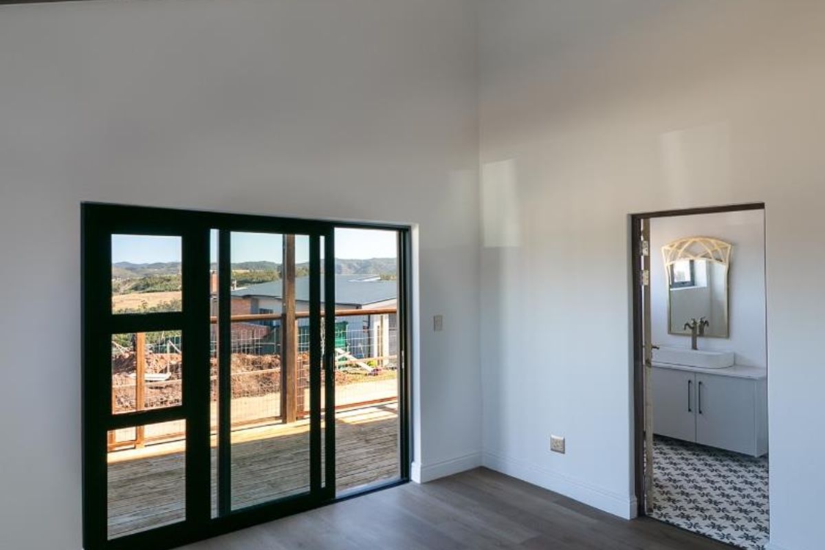 A bright bedroom featuring exposed timber ceiling beams, wood-look flooring, and sliding glass doors leading to an outdoor deck, with an adjoining en-suite bathroom.