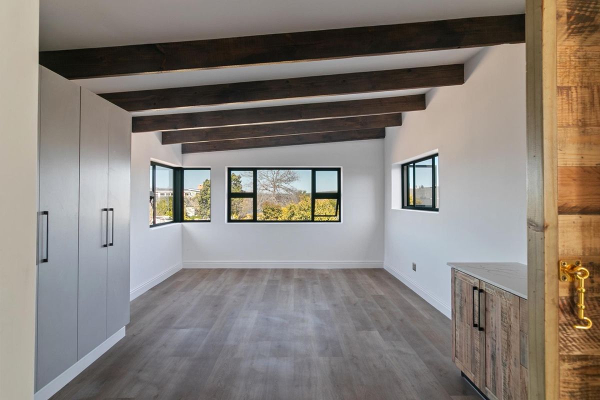 A bright, modern living space featuring exposed timber ceiling beams, sleek grey cabinetry, and large windows offering natural light and scenic views.