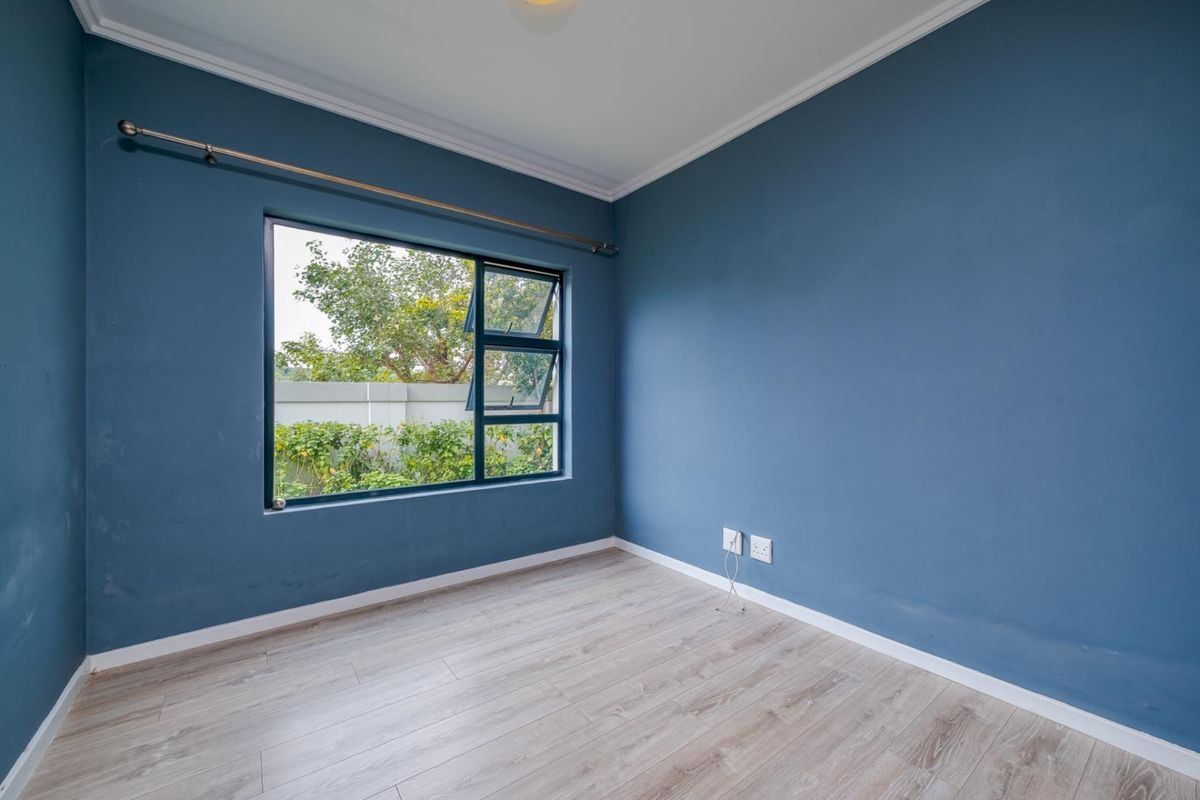 A bright, empty bedroom featuring blue painted walls, light wood-look laminate flooring, and a large window with a view of the garden.