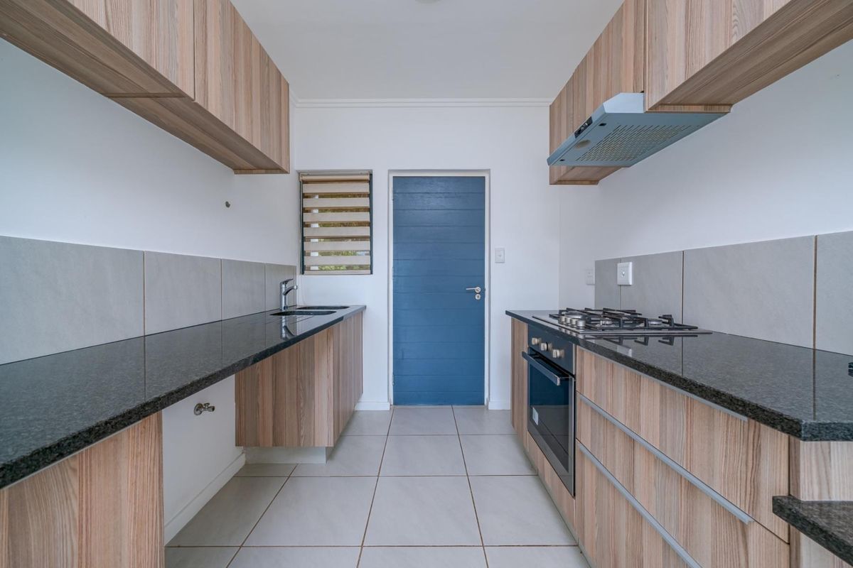 Modern galley-style kitchen featuring wood-grain cabinetry, dark granite countertops, a built-in gas hob, oven, and extractor fan with tiled flooring.