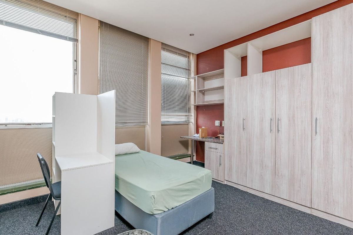 A bedroom featuring built-in light wood-grain cabinetry, a desk area, and large windows with fitted blinds.