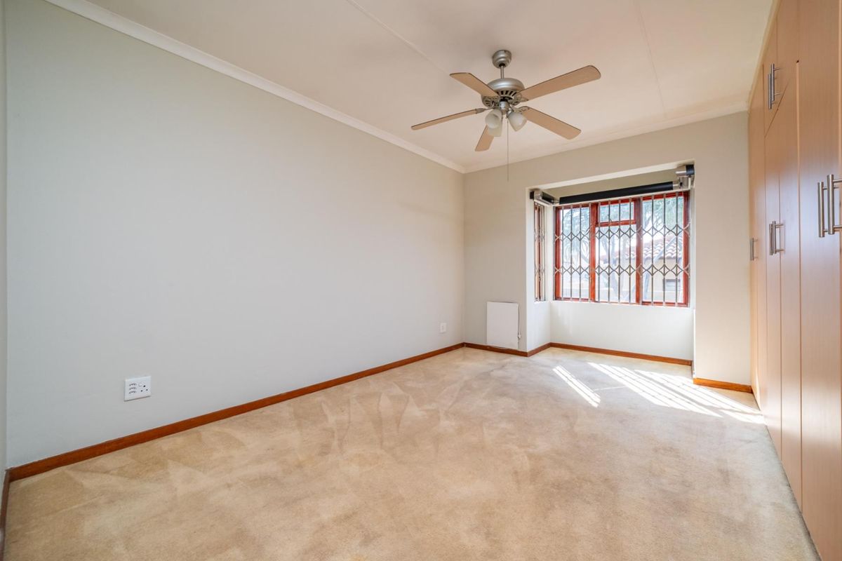 A bright, empty bedroom featuring neutral walls, built-in wooden cupboards, a ceiling fan, and a window fitted with a security gate.