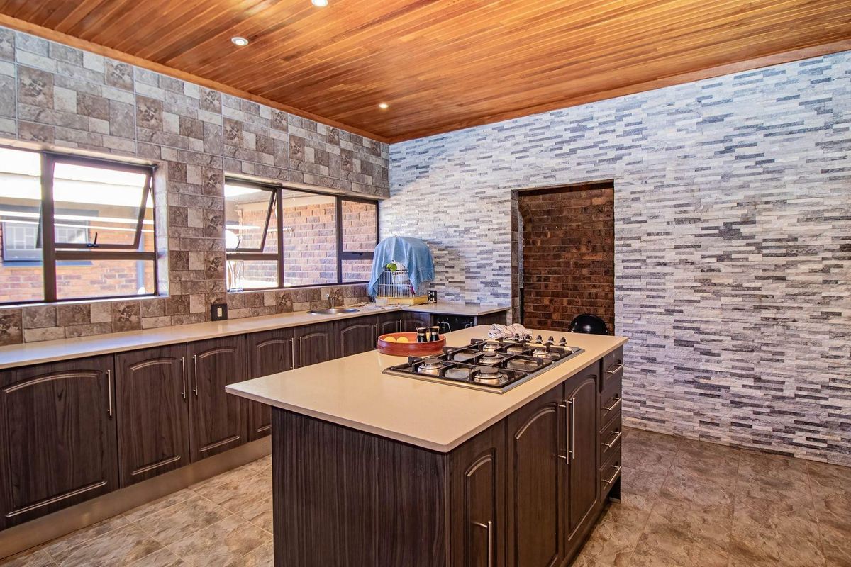 Spacious kitchen featuring a central island with a built-in gas hob, dark wood cabinetry, stone-look wall tiles, and a warm timber-paneled ceiling.