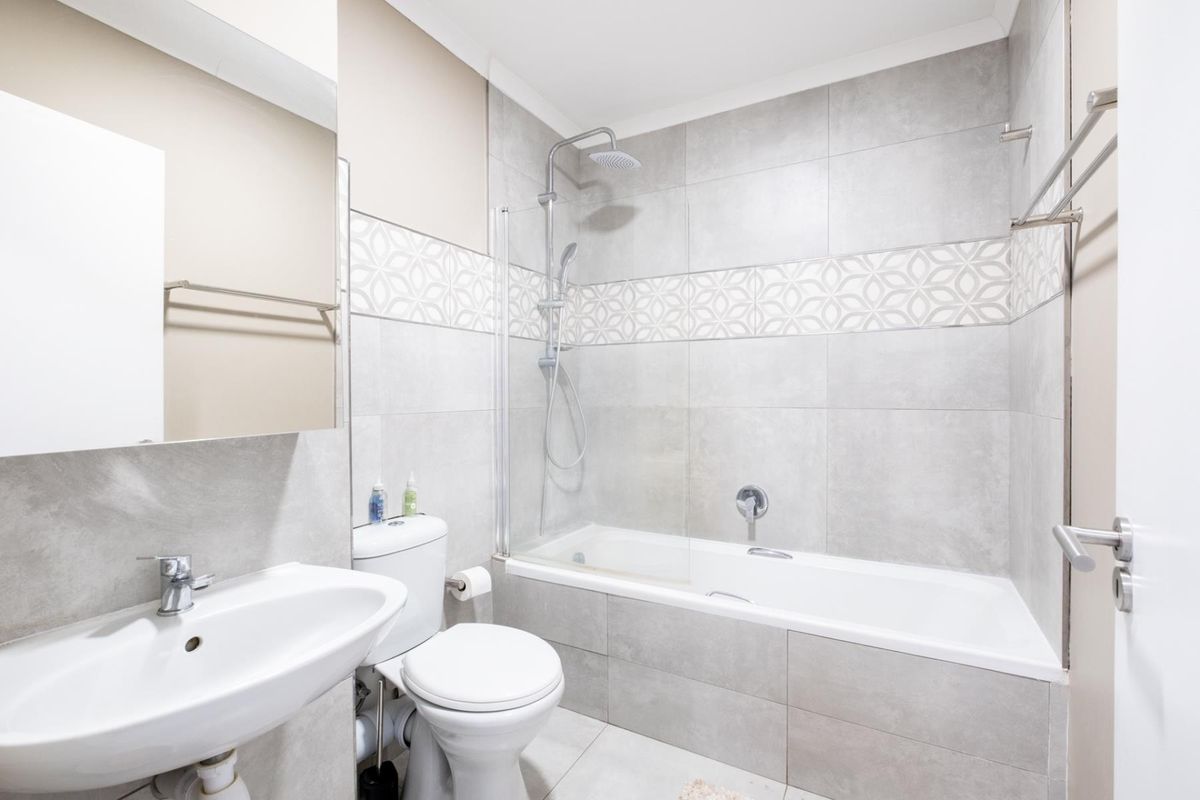 Modern bathroom featuring a bathtub with a glass shower screen, wall-mounted shower head, pedestal sink, and neutral grey wall tiling.