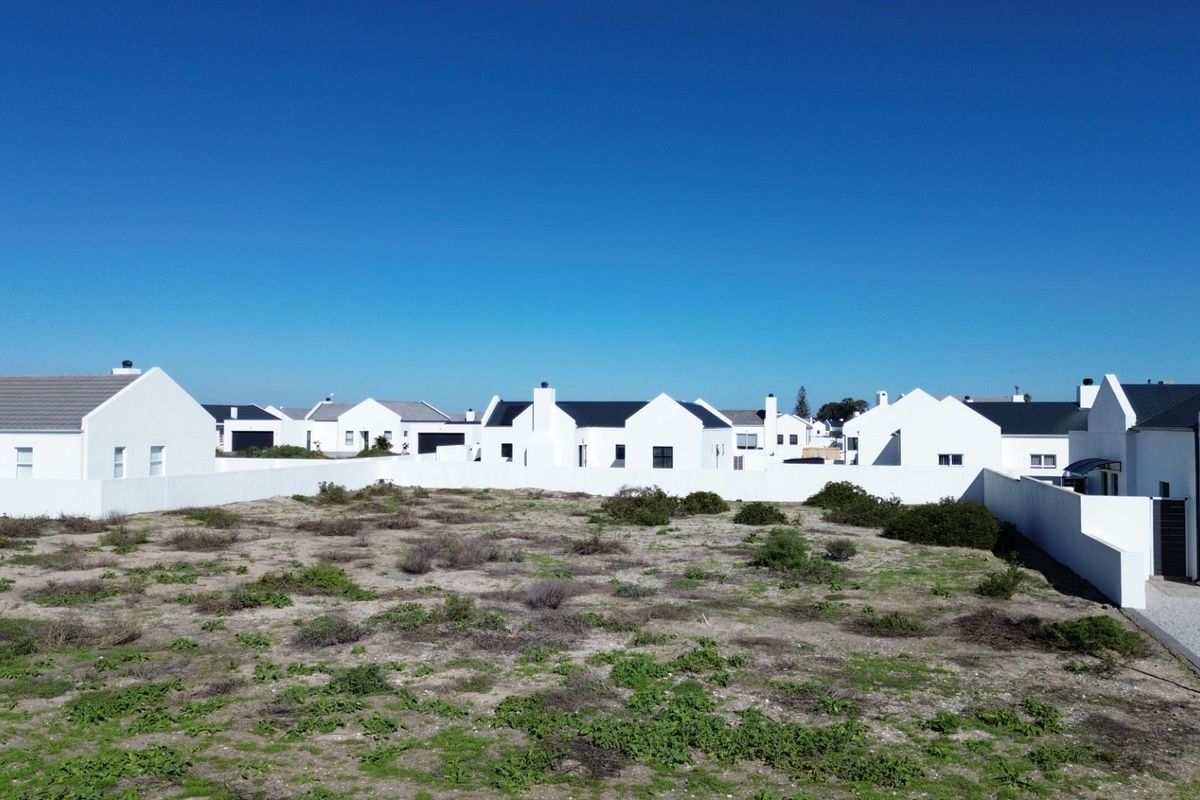 A vacant residential plot of land surrounded by established white-walled homes in a quiet neighborhood setting.