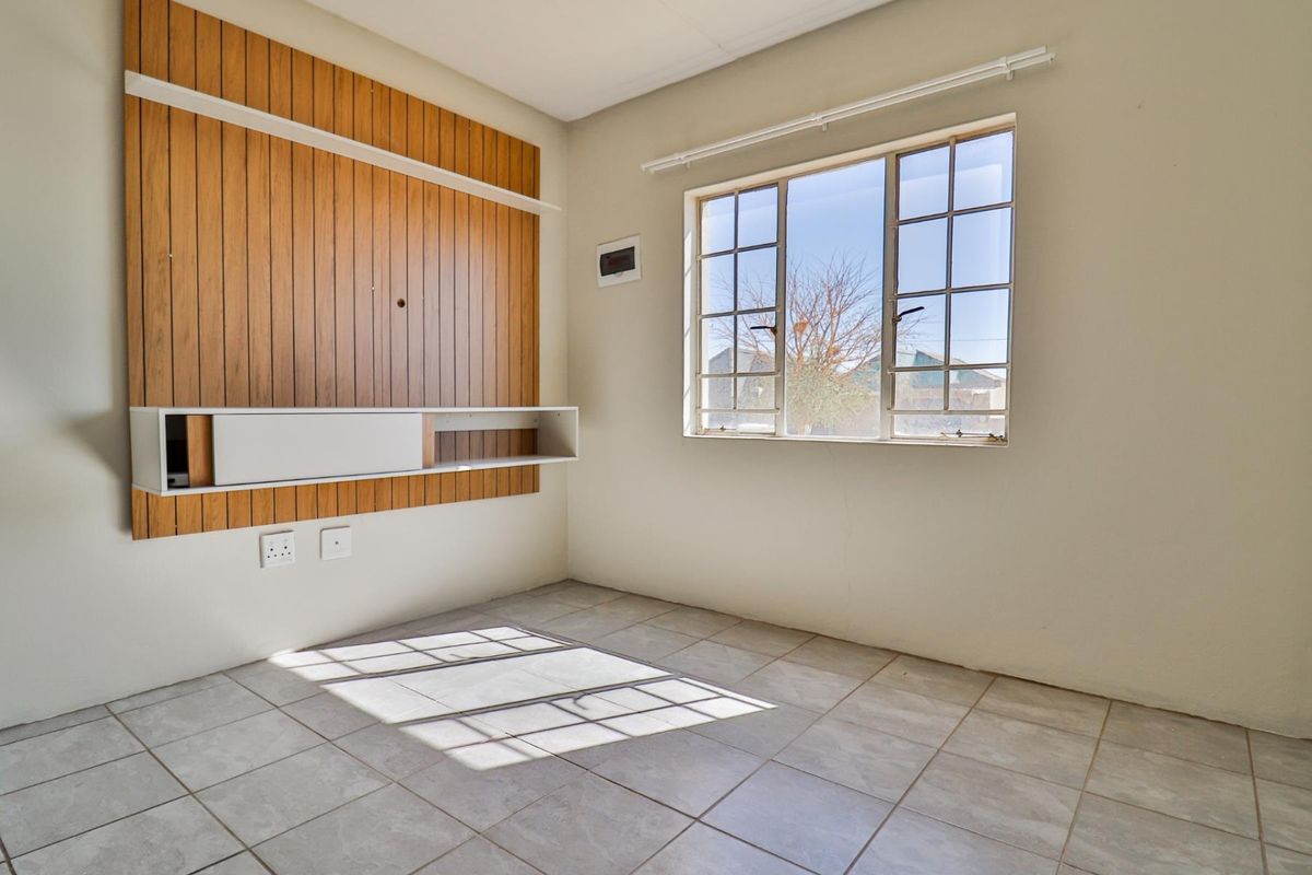 An empty living room featuring tiled flooring, a large grid-pattern window, and a built-in wooden wall feature with a floating shelf.