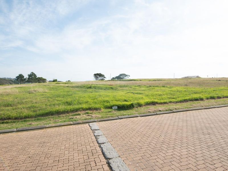 A vacant residential plot of land featuring level grass terrain, accessible from a paved street with established kerbing.
