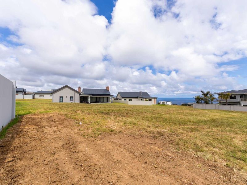 A vacant residential plot featuring a grassed landscape, boundary wall, and views of neighboring properties and the distant coastline.