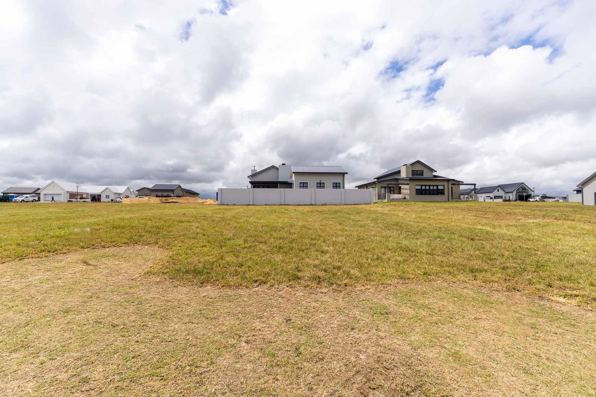 A wide-angle view of a vacant residential plot featuring a grass lawn and a boundary wall, with neighboring houses visible in the background.