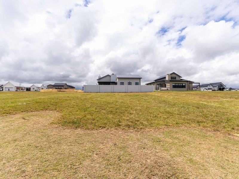 A wide-angle view of a vacant residential plot featuring a grass lawn and a boundary wall, with neighboring houses visible in the background.