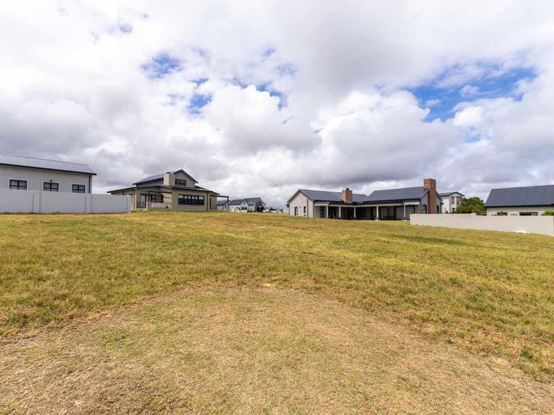 A wide-angle view of a vacant residential plot featuring a grass lawn, surrounded by established modern homes and boundary walls.