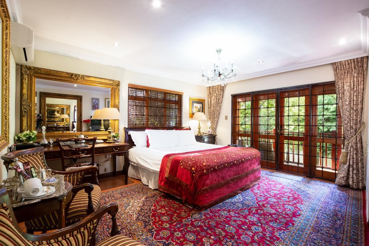 A well-appointed hotel bedroom featuring a large bed, ornate gold-framed mirror, recessed ceiling lighting, and wooden French doors leading to an outdoor balcony.