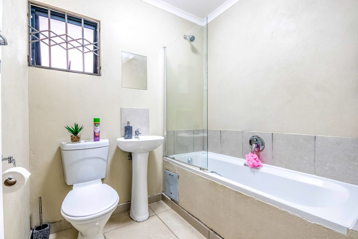 A functional bathroom featuring a pedestal basin, toilet, and a bathtub with a glass shower screen and security-barred window.