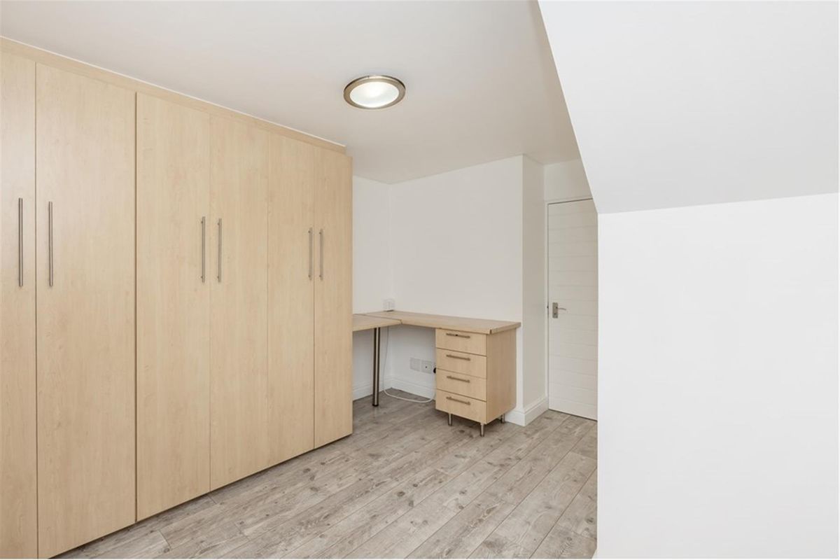 A functional bedroom featuring light wood-grain built-in wardrobes and a matching integrated desk unit with drawers, set against neutral walls and wood-look flooring.