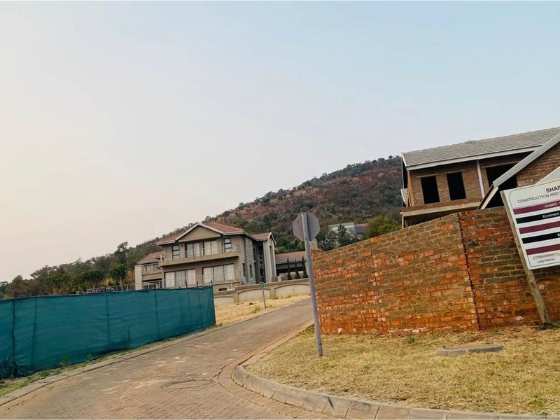 A view of a residential street in a developing estate, featuring a brick boundary wall, construction signage, and surrounding hillside landscape.