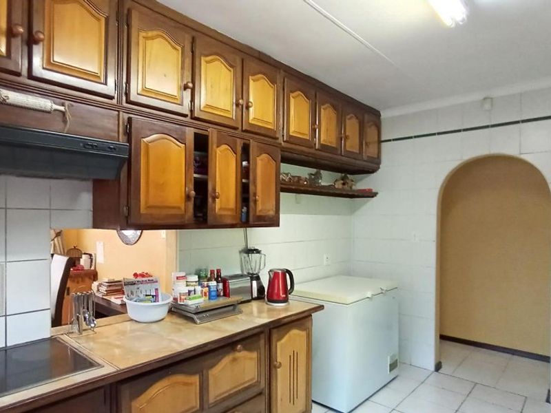 A dated kitchen featuring traditional wooden cabinetry, tiled splashbacks, and tiled flooring, with an arched doorway leading to an adjacent room.