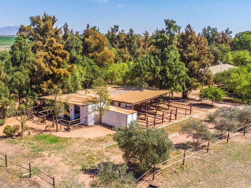 Aerial view of a farm property featuring stable structures with corrugated metal roofing, surrounded by mature trees and fenced paddocks.