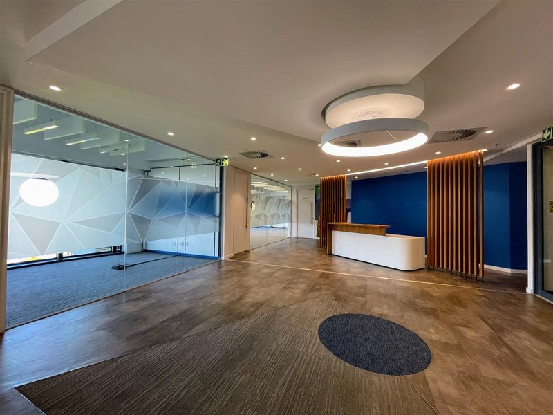 Modern office reception area featuring a sleek reception desk, glass-partitioned offices, contemporary lighting, and a combination of carpet and vinyl flooring.