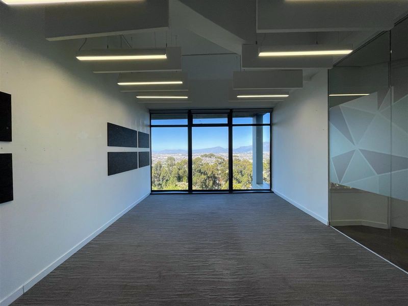 A bright, empty office space featuring modern suspended lighting, floor-to-ceiling windows with expansive mountain views, and a glass partition wall.