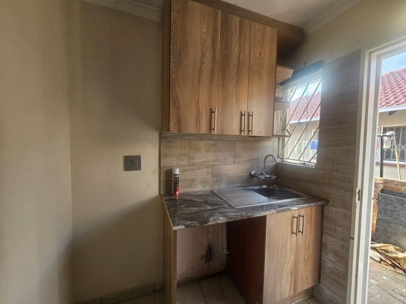 A compact scullery area featuring wood-look cabinetry, a stainless steel sink, and tiled splashbacks, with direct access to an outdoor area.