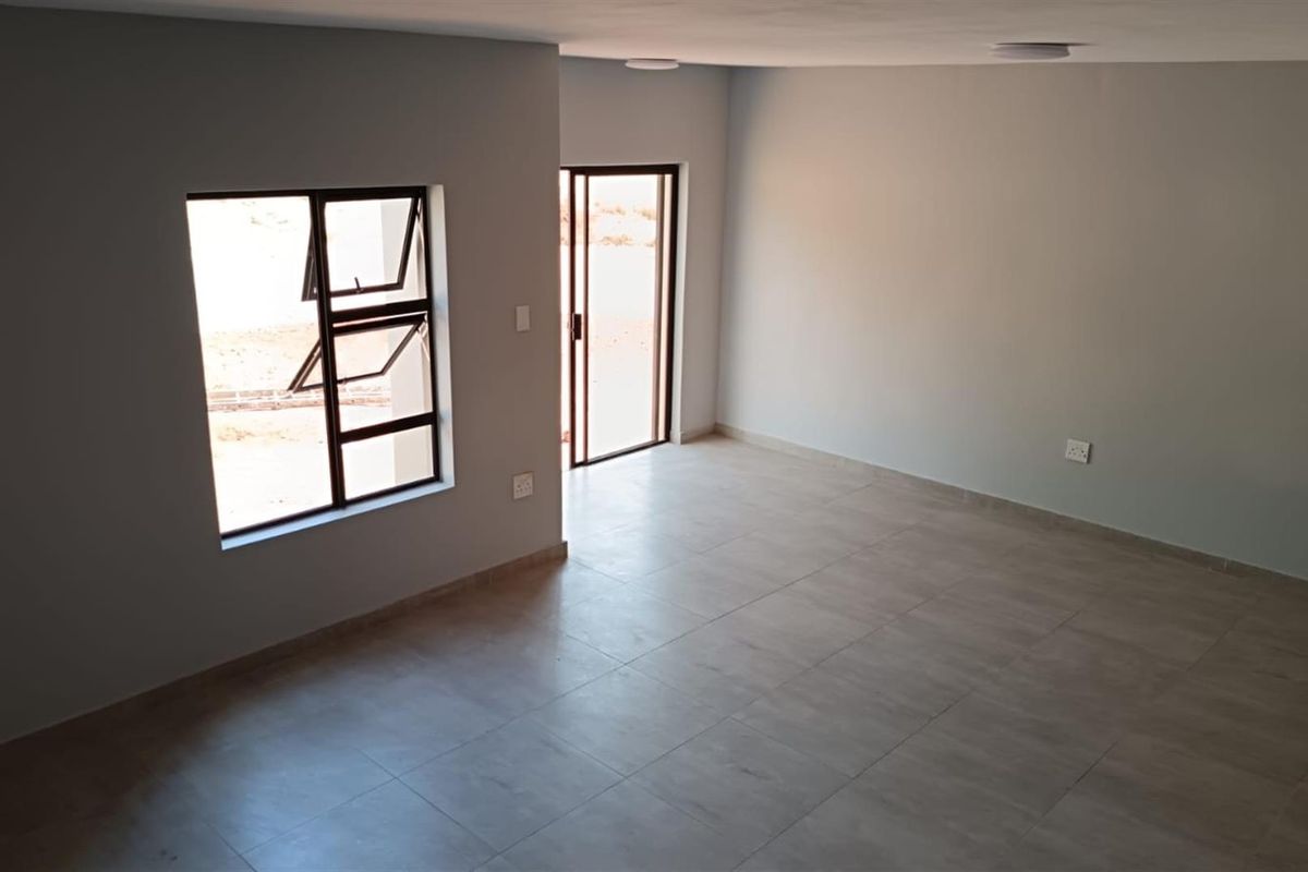 A bright, empty living room featuring neutral tiled flooring, modern aluminium window frames, and a sliding glass door leading to the exterior.