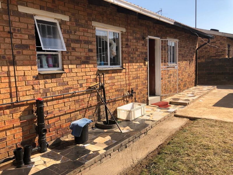Rear exterior view of a face brick house featuring a tiled patio area, security window bars, and exposed external plumbing infrastructure.