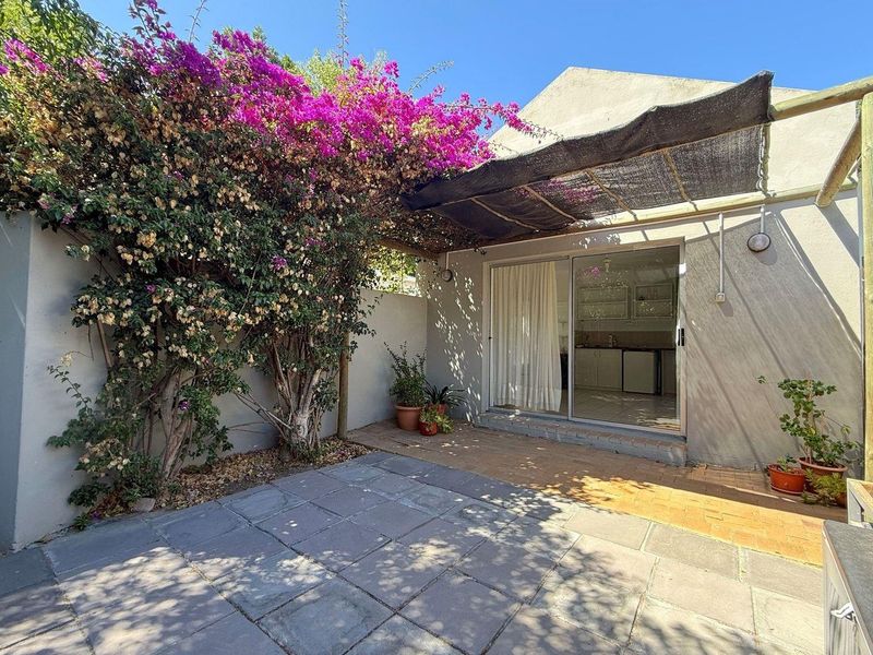 A paved outdoor patio area featuring a shade cloth pergola, established bougainvillea, and direct access to the interior through a sliding glass door.