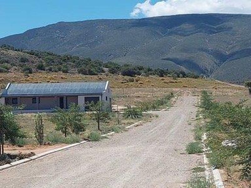 A view of a rural property featuring a single-story house situated along a gravel access road with scenic mountain views in the background.