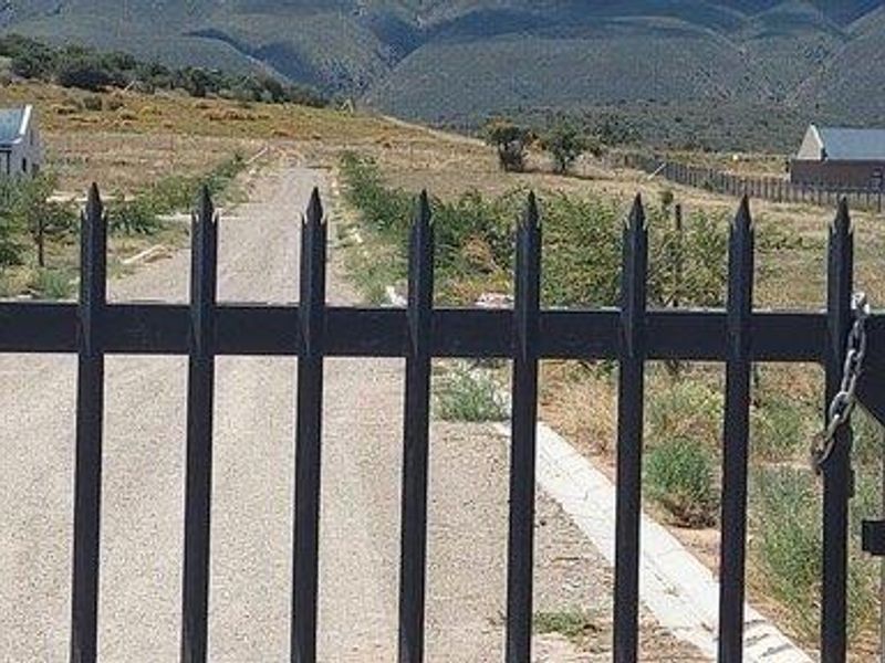 A view through a metal security gate looking down a gravel driveway towards rural buildings and mountain scenery.