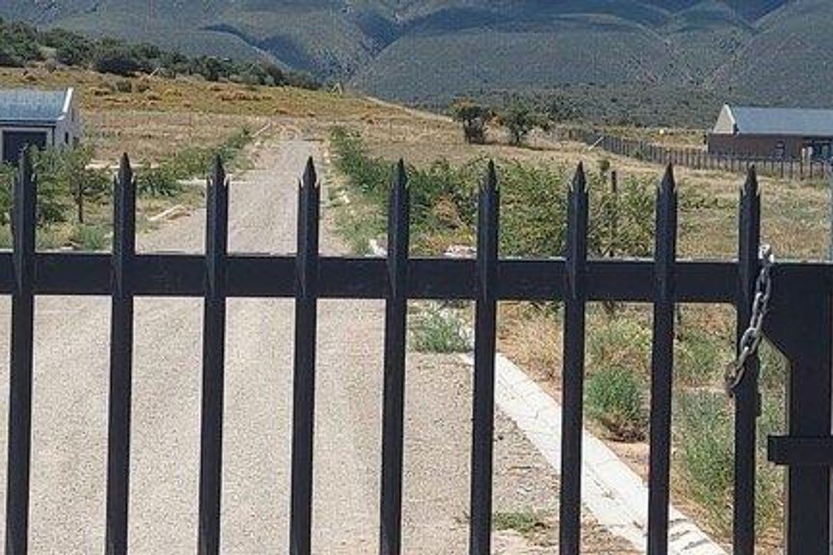 A view through a metal security gate looking down a gravel driveway towards rural buildings and mountain scenery.