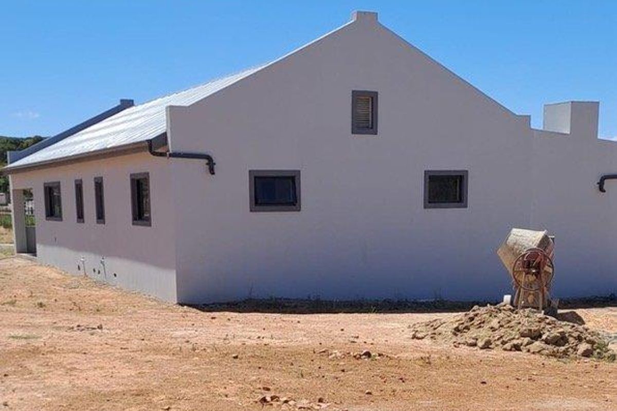 Exterior view of a modern house with a gabled roof, featuring clean rendered walls and dark window frames, set on an undeveloped plot.