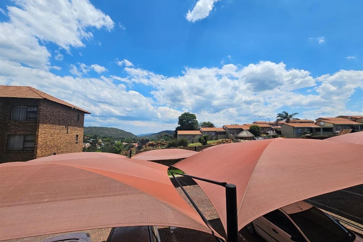 An elevated view of a residential complex parking area featuring shade sail structures and paved driveway surfaces with surrounding hillside views.