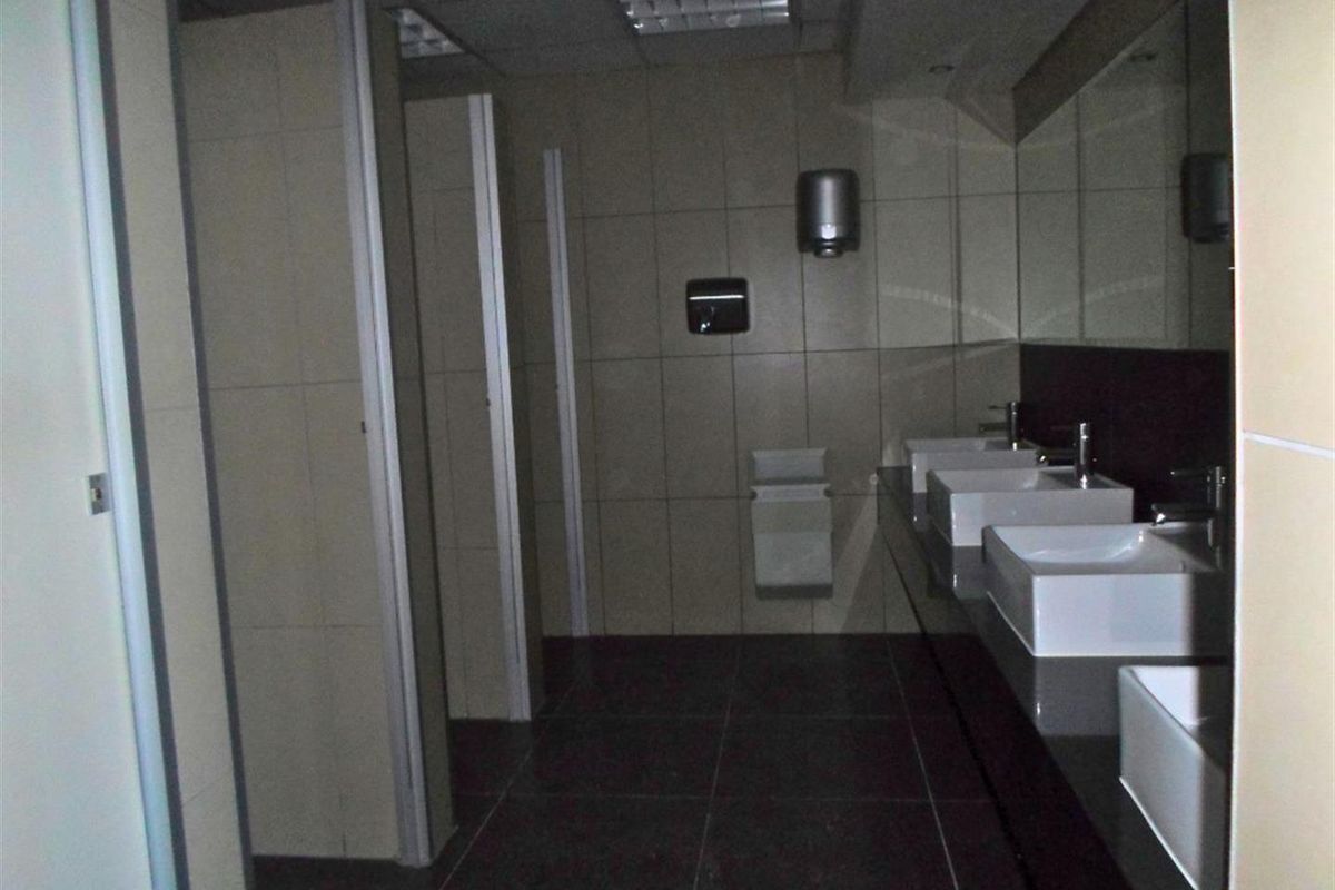 Commercial bathroom facility featuring multiple washbasins, a large vanity mirror, tiled surfaces, and private toilet cubicles.